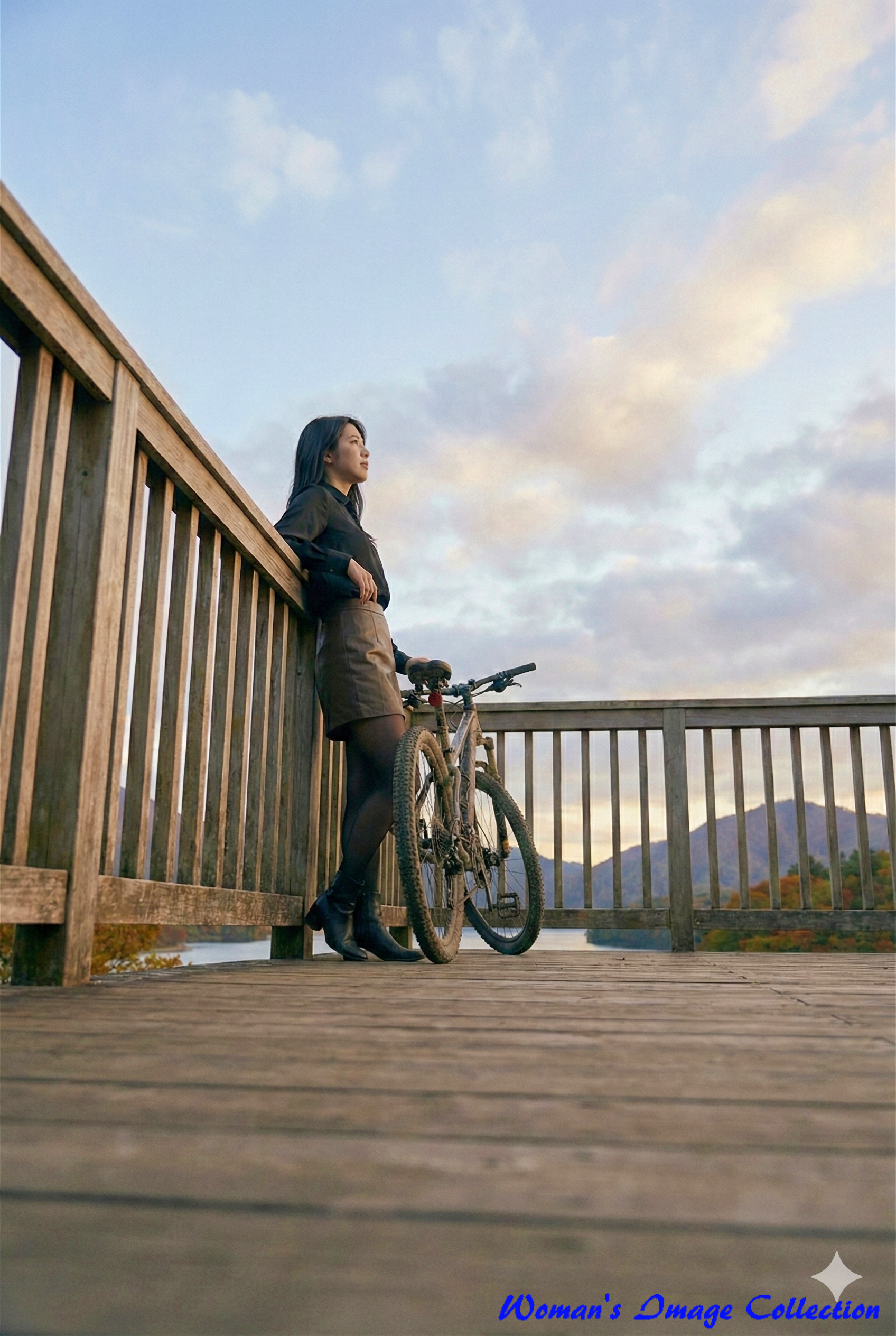 夕暮れの湖畔のデッキで手すりに寄りかかり、遠くを見つめる女性とマウンテンバイク。