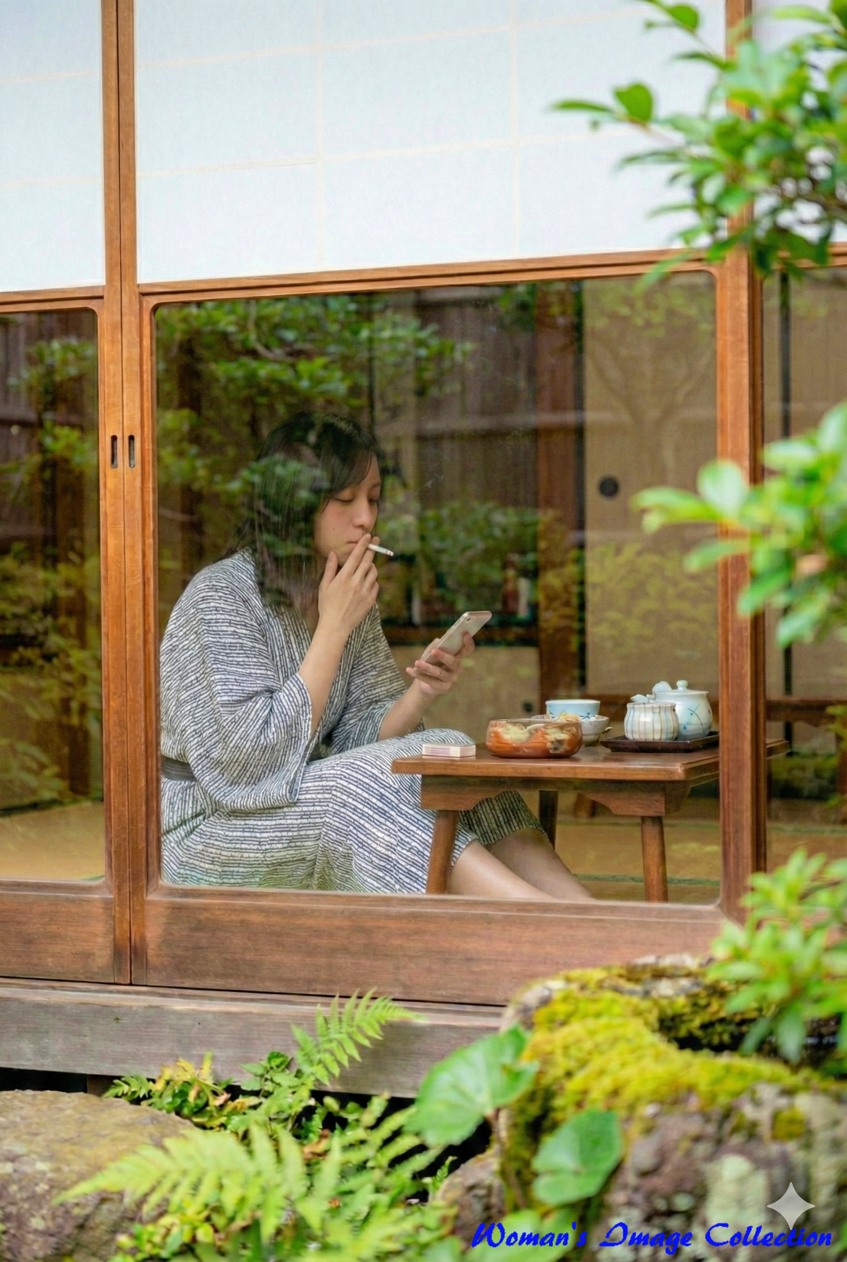 庭先のガラス戸越しに撮影された、和室でタバコを吸う浴衣姿の女性