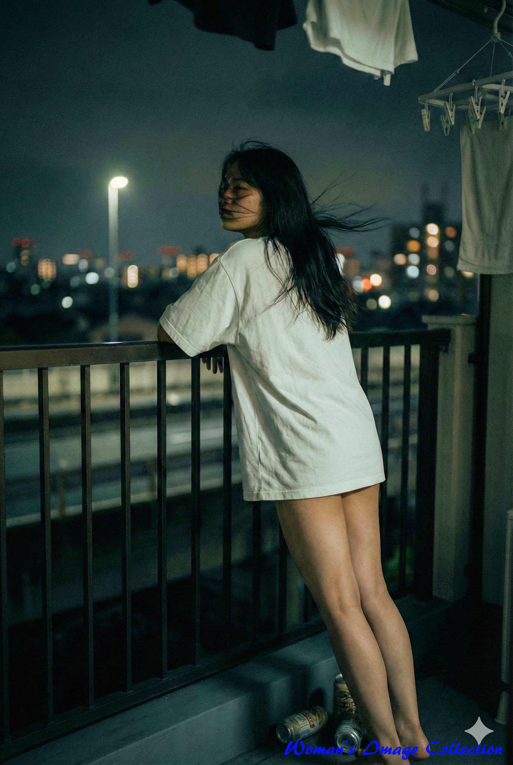 足元に空き缶が置かれた夜のバルコニー。暗闇の中で白いTシャツが浮かび上がり、遠くの街明かりが丸くボケている様子。